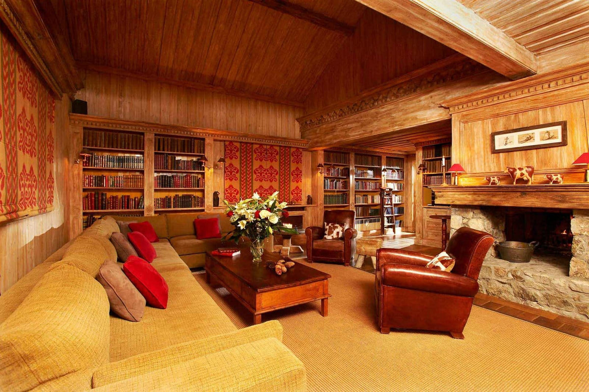 A cozy, rustic wood-paneled living room with a large mustard yellow sofa, red accent pillows, a dark wood coffee table with flowers, a brown leather armchair, built-in bookshelves, and a stone fireplace.