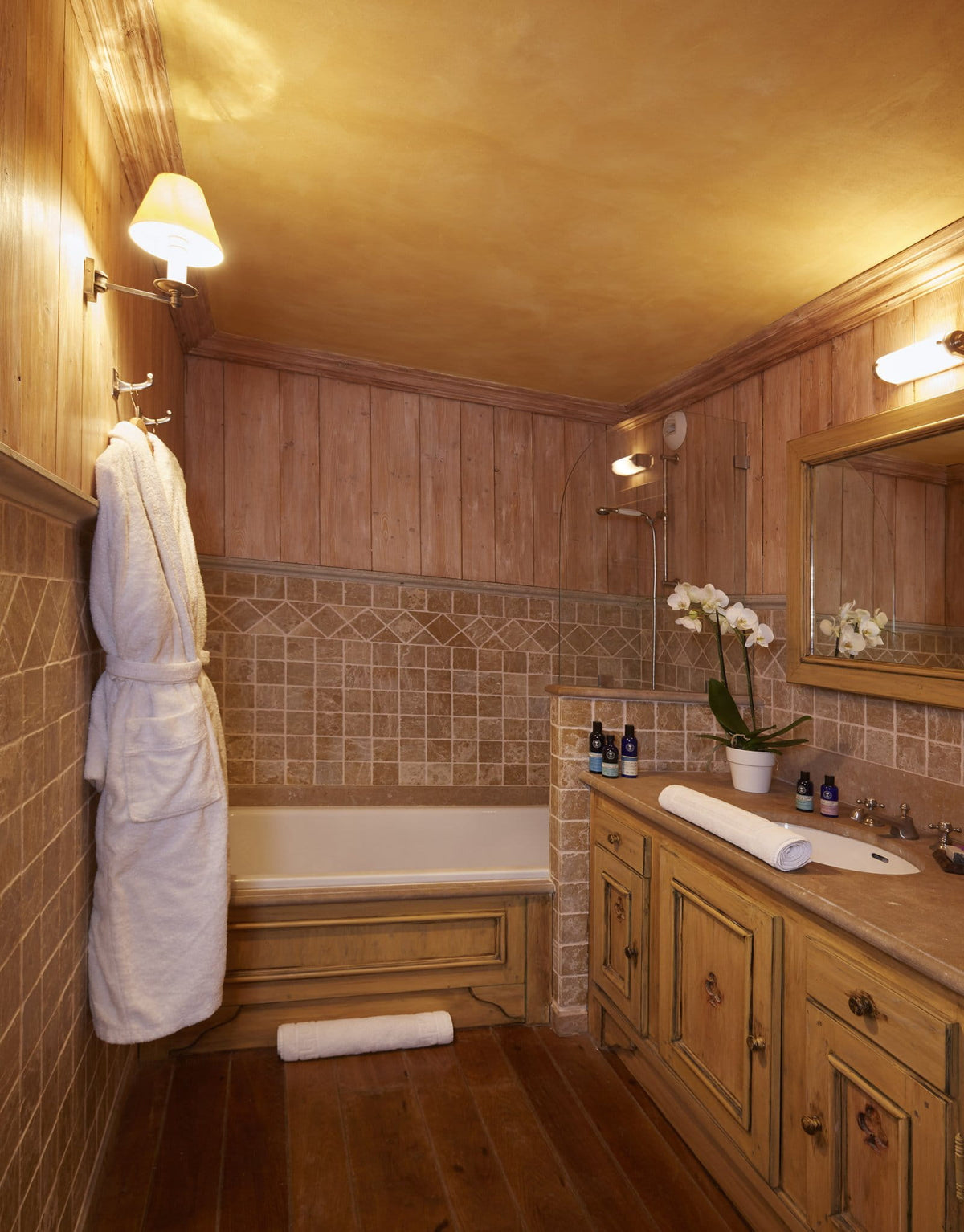 A rustic bathroom interior with warm wood-paneled walls, a built-in bathtub, a matching vanity with a mirror, and a hanging white bathrobe.