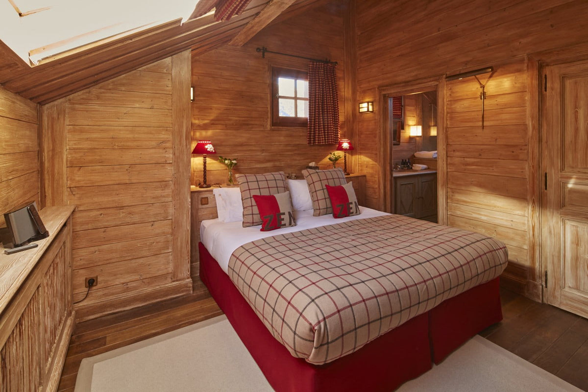 A cozy mountain chalet bedroom featuring a large bed with a beige and red plaid quilt, red bed skirt, and matching accent pillows, set against warm wood-paneled walls.