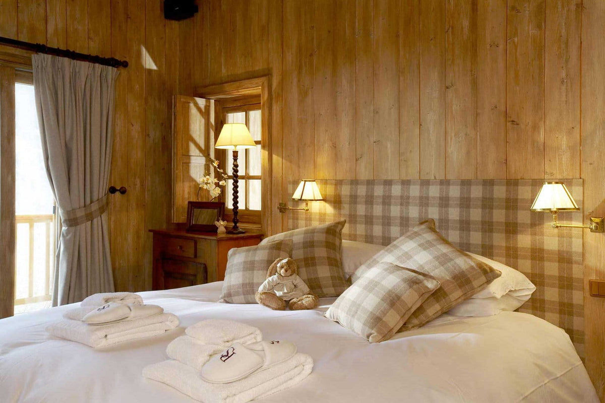 A cozy bedroom interior with a large bed dressed in white linens, plaid pillows, and folded white towels, set against warm wood-paneled walls with bedside lamps and a window.