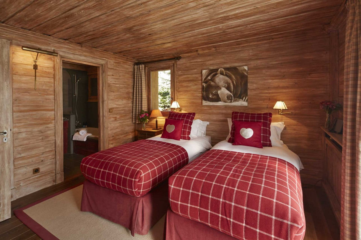 A cozy mountain chalet bedroom with two twin beds dressed in red and white plaid blankets and matching pillows, set against warm wood-paneled walls and ceiling.