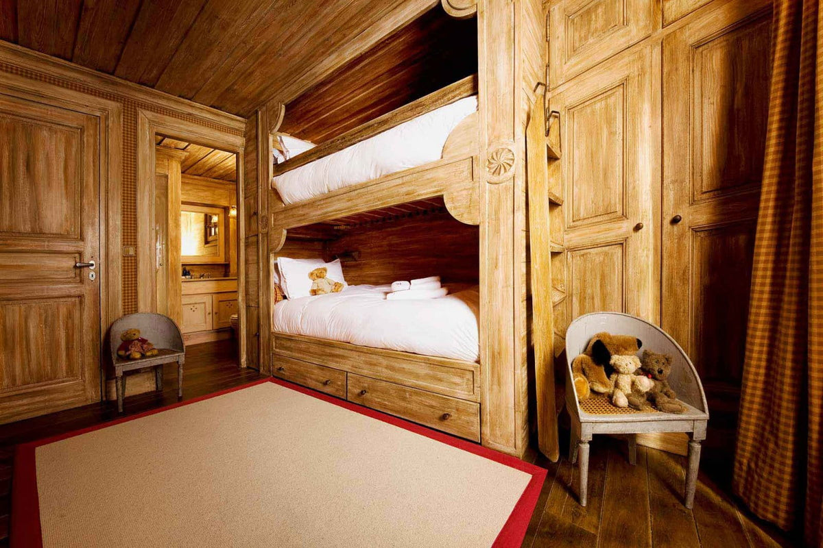 A rustic wooden bedroom featuring a built-in wooden bunk bed with white bedding, a cream rug with a red border, a white chair with stuffed toys, and warm wood-paneled walls and ceiling.