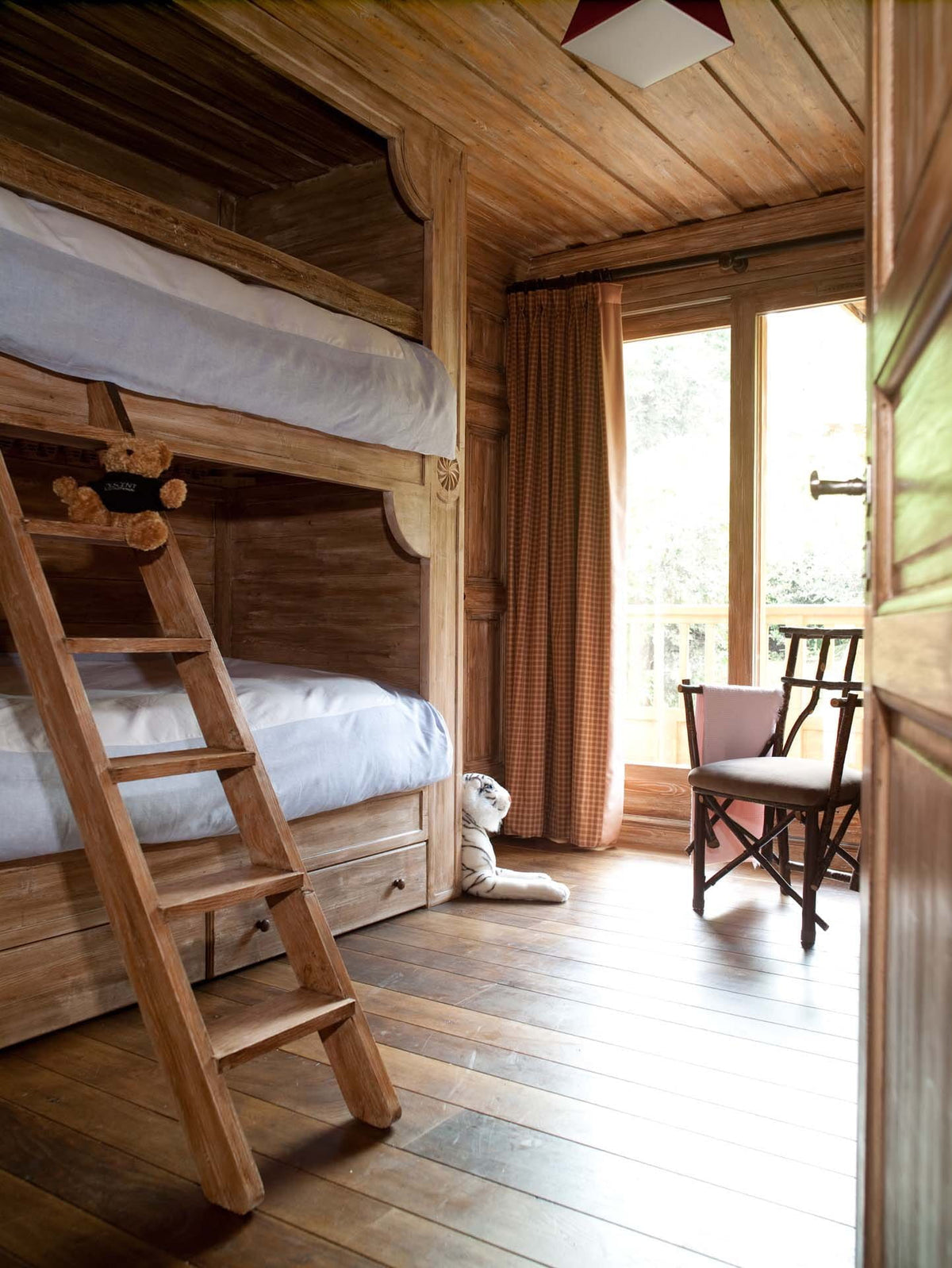 A rustic wooden bunk bed with a wooden ladder in a cozy cabin bedroom, featuring a window with orange curtains, a wooden chair, and a white stuffed animal on the floor.
