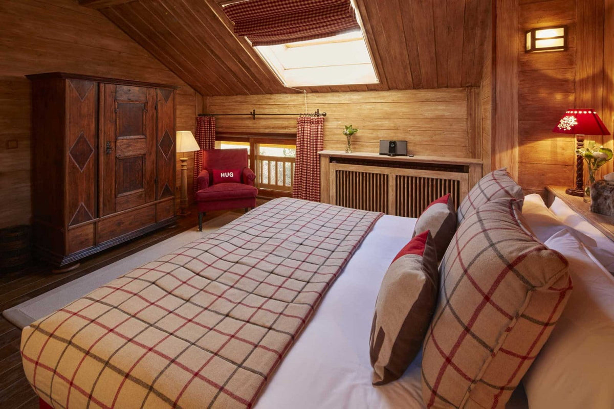 A cozy rustic bedroom with a large bed dressed in beige and red plaid bedding, set in a wood-paneled attic room with a red armchair, wooden wardrobe, and skylight.