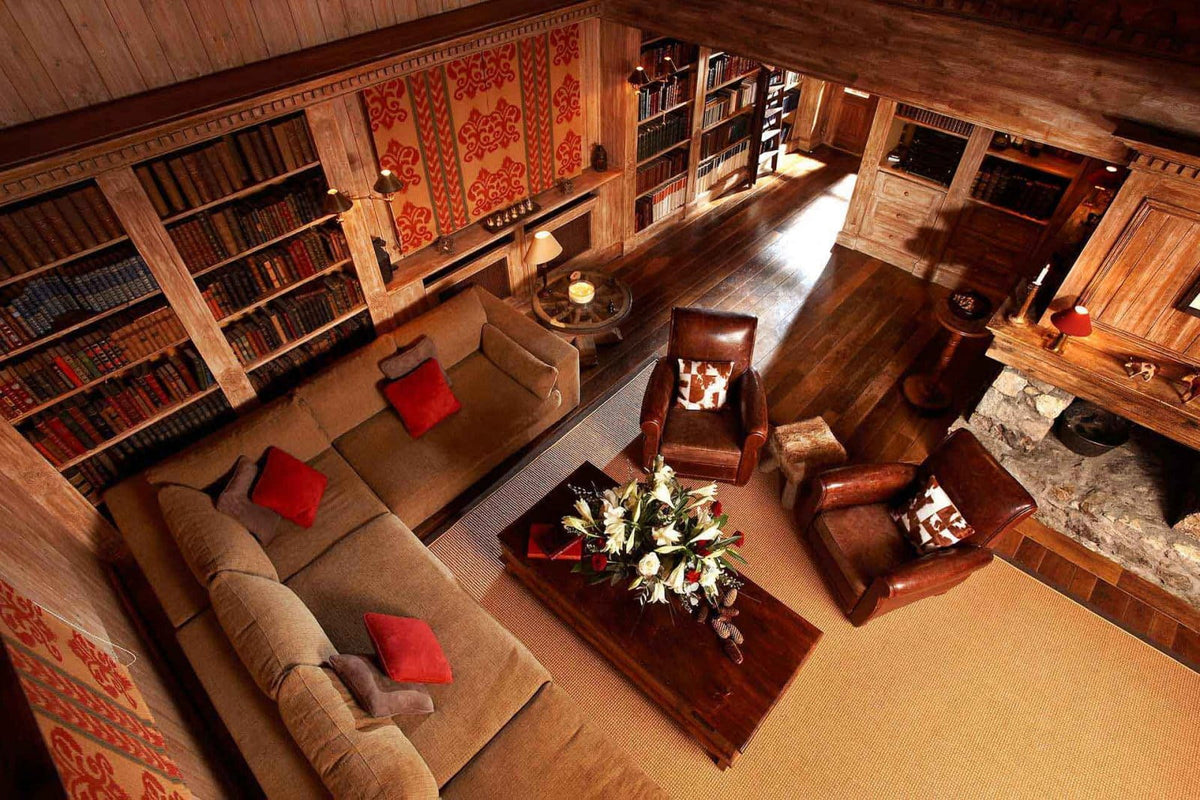 A cozy, rustic mountain-style living room with a large beige sectional sofa, two brown leather armchairs, a dark wood coffee table with a floral arrangement, and extensive built-in wooden bookshelves along the walls.