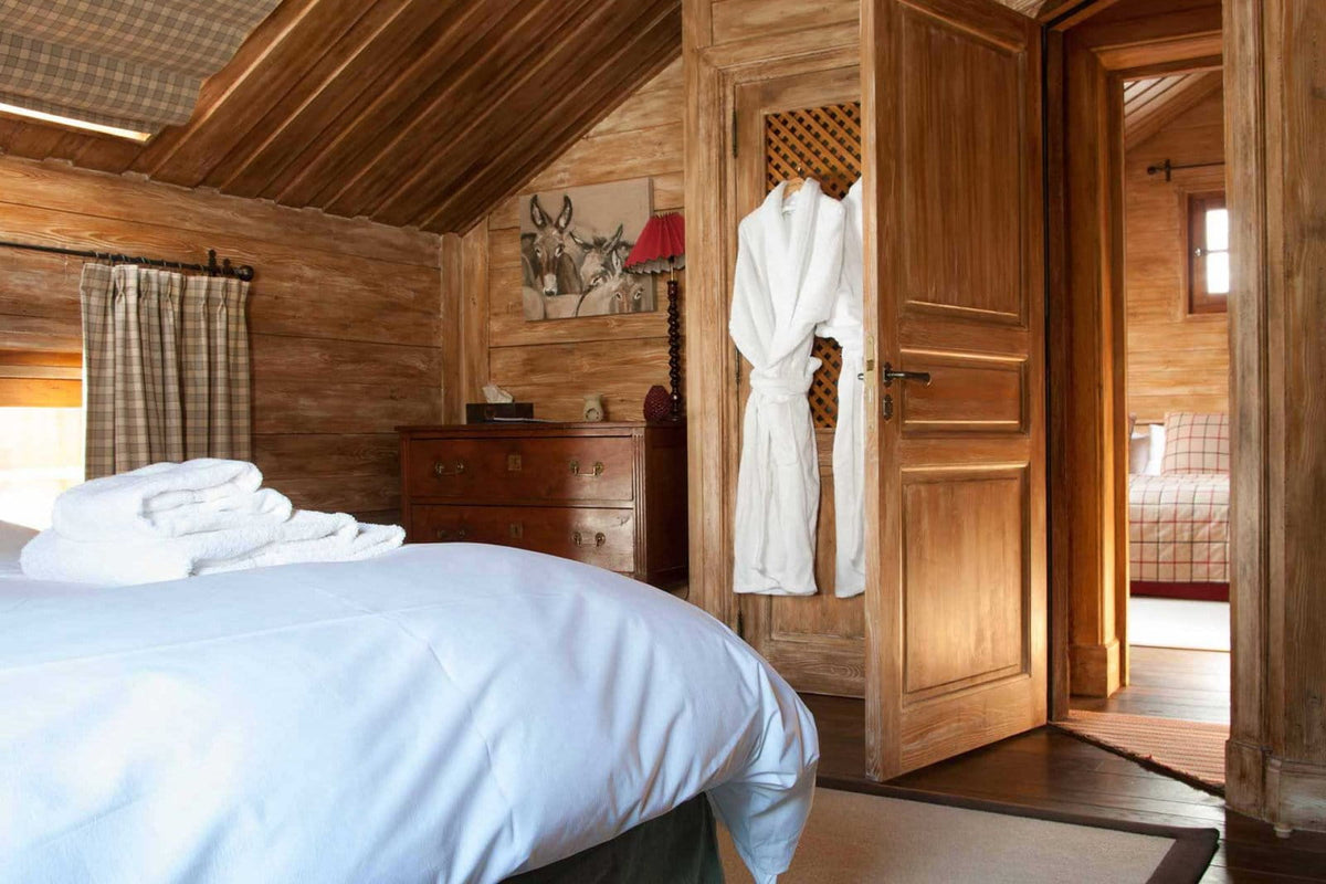 A rustic bedroom with wood-paneled walls and ceiling, featuring a bed with white bedding in the foreground, a wooden dresser, and white bathrobes hanging on a wooden door.