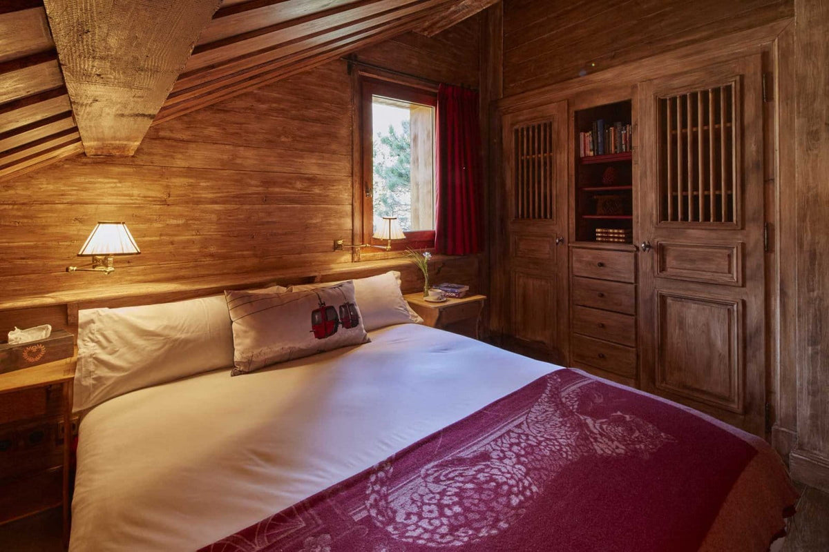 A cozy rustic bedroom with a wooden bed dressed in white linens and a deep red patterned runner, set against warm wood-paneled walls with a window, bedside lamps, and a wooden cabinet.