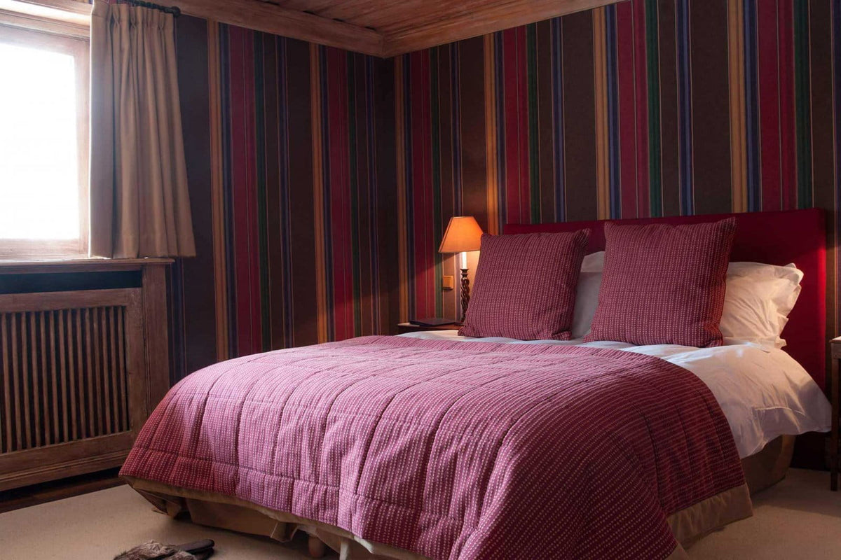 A cozy bedroom scene with a bed dressed in a textured magenta quilt and matching pillows, set against multicolored vertically striped wallpaper. A lit bedside lamp and a window with curtains are visible in the room.