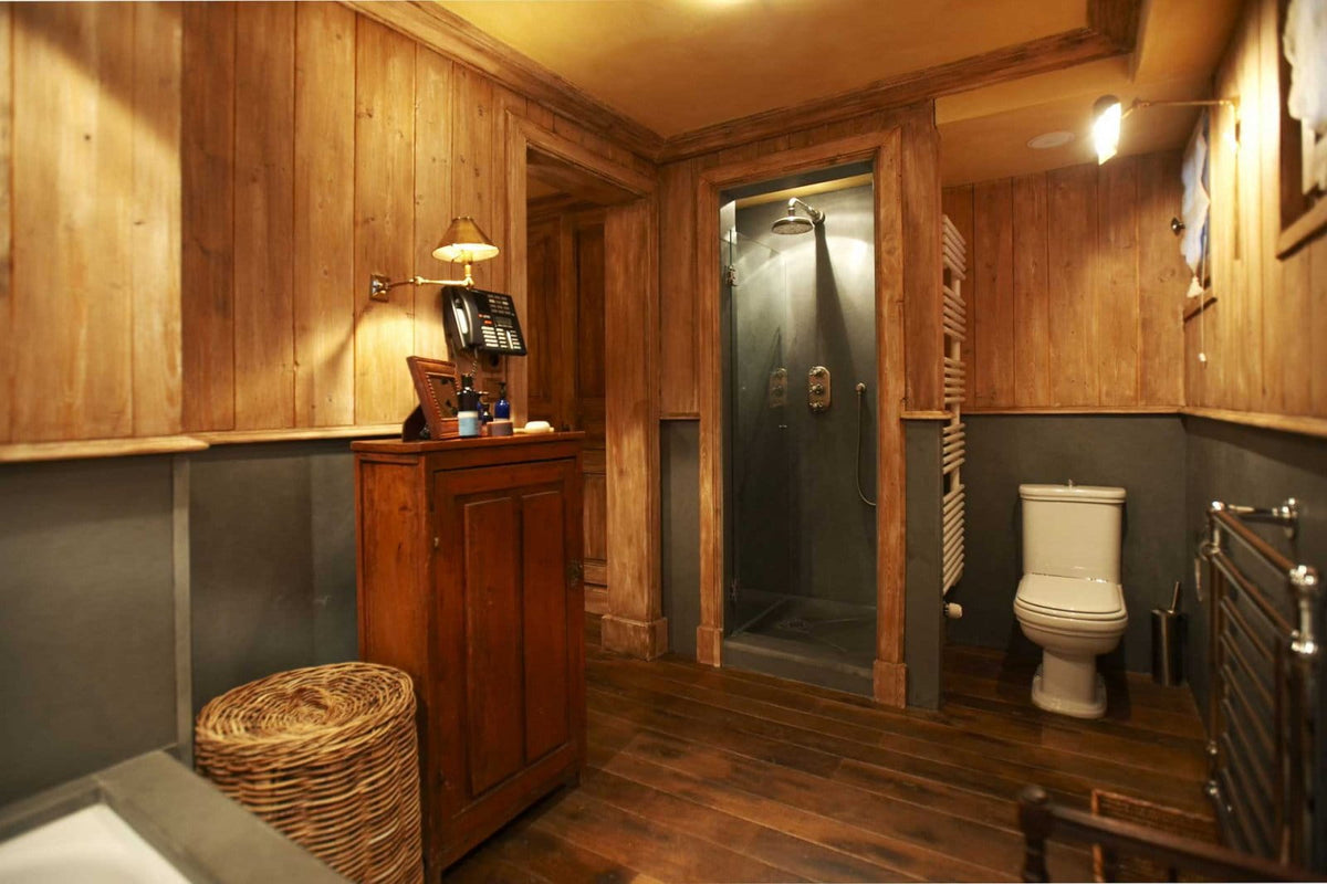 A rustic wooden bathroom interior with wood-paneled walls, a dark wood vanity cabinet, a wicker basket, a white toilet, and a glass shower enclosure.