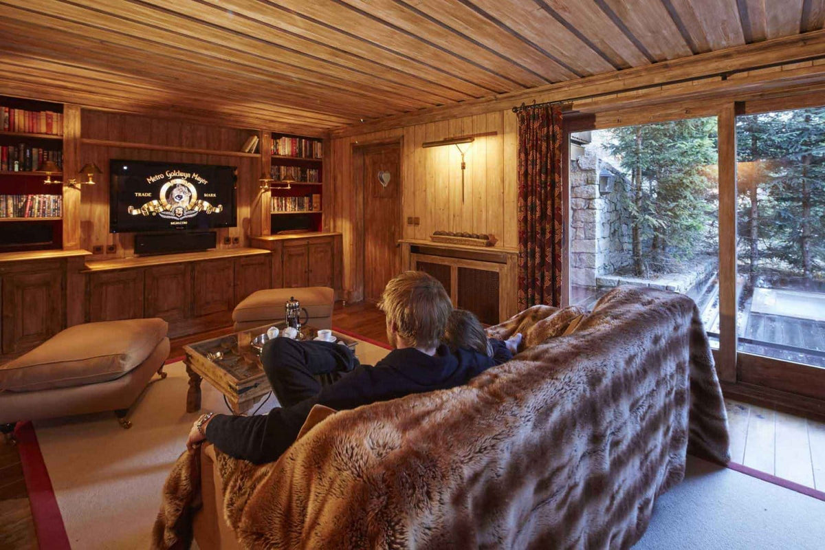 A cozy, rustic wood-paneled living room with a large tufted brown sofa in the foreground, a TV mounted on a wooden wall, and two people sitting on the sofa facing the screen. Large windows on the right show a wooded outdoor setting.