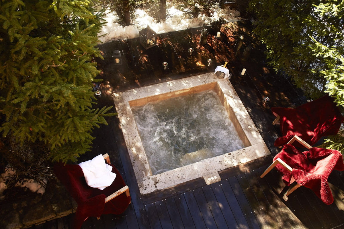 An overhead view of a small outdoor hot spring or bubbling pool on a dark wooden deck, surrounded by green trees and two red lounge chairs with white towels.