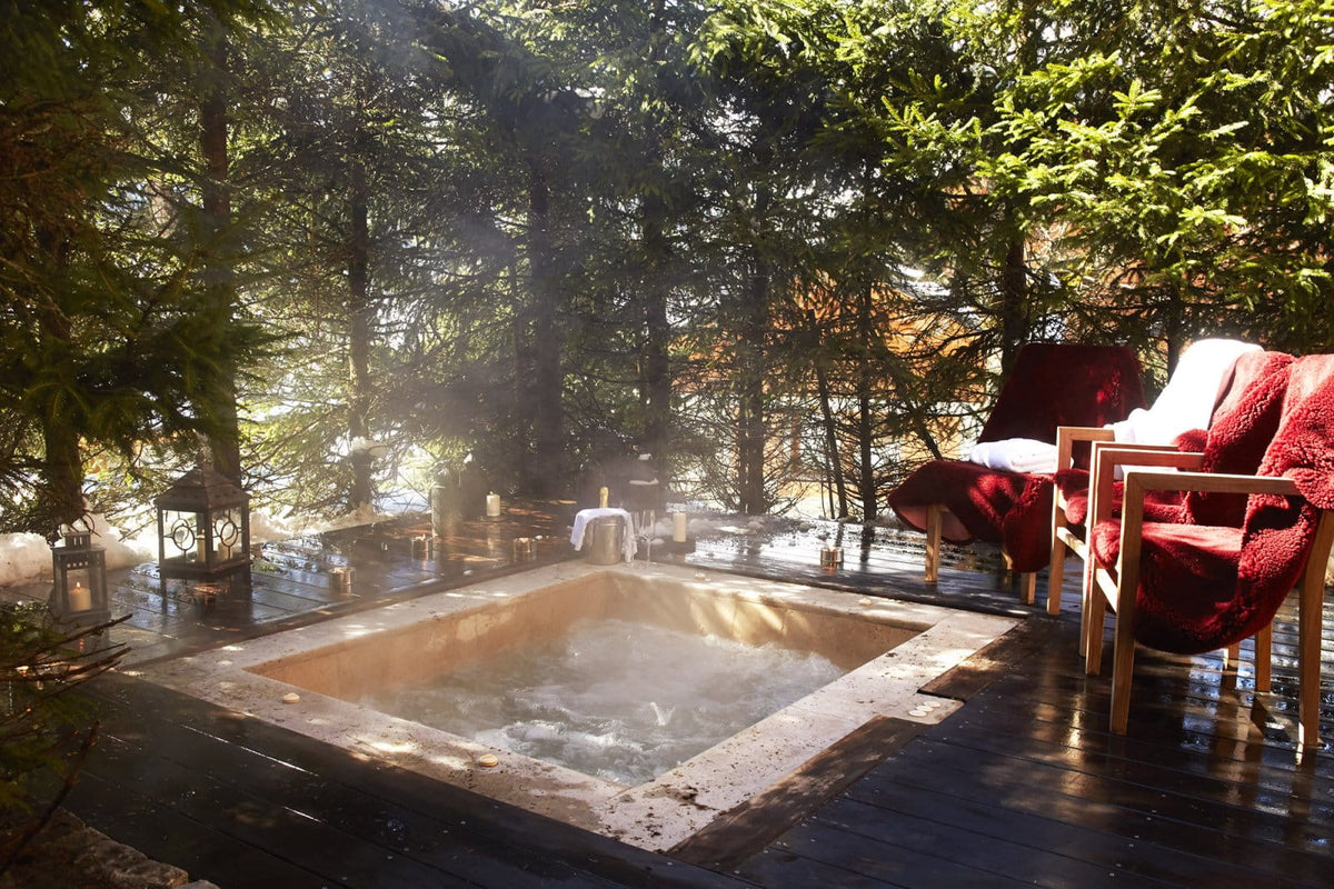 An outdoor hot spring or small pool on a wooden deck surrounded by evergreen trees, with two red lounge chairs draped in red blankets on the right.
