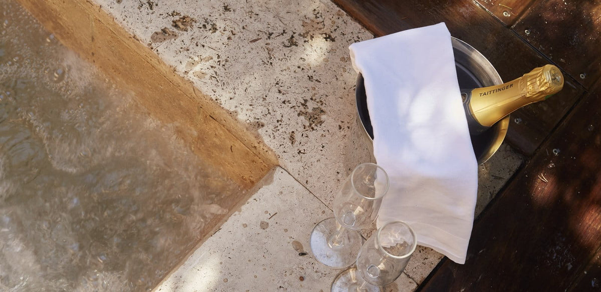 A bottle of champagne in an ice bucket with a white napkin, alongside two clear champagne flutes, set on a dark wooden table next to a stone floor.