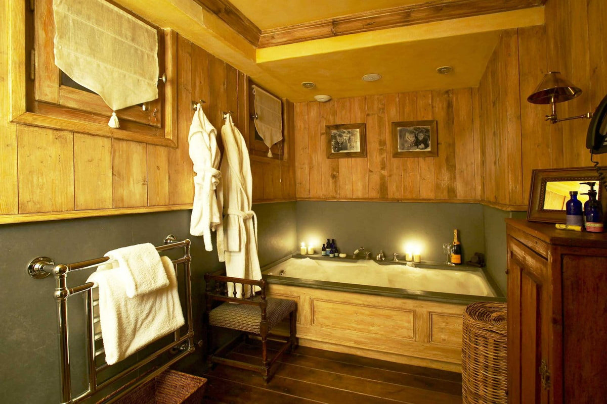 A rustic wooden bathroom interior with a built-in bathtub, warm wood paneling, hanging white robes, a towel rack, and ambient lighting.