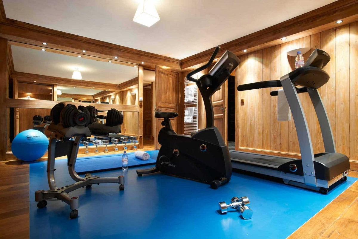 A home gym interior with multiple exercise machines: a treadmill, elliptical, free weight rack with dumbbells, and a blue exercise ball on a blue mat, set against wood-paneled walls.