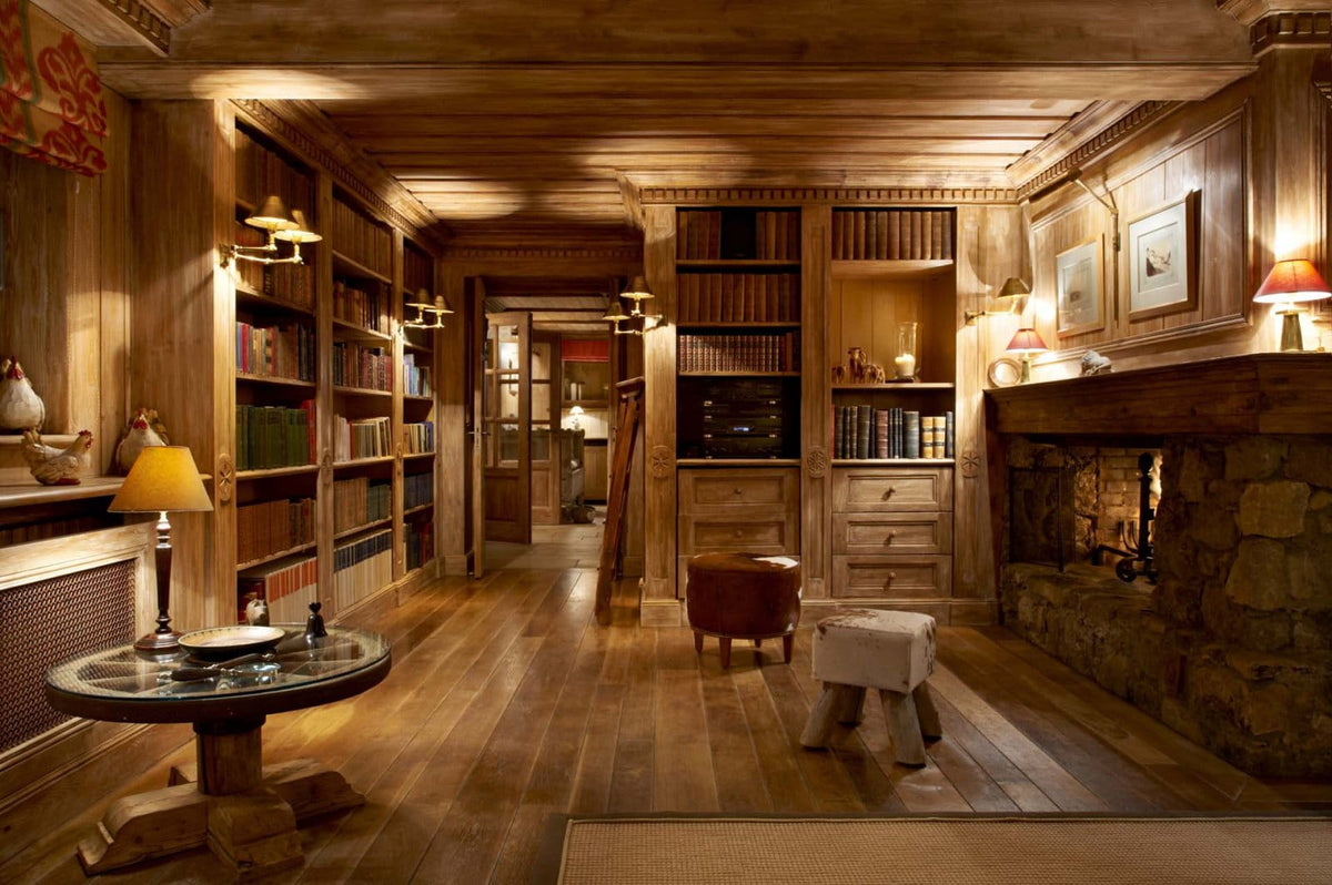 A warm, rustic library or study with dark wood walls, bookshelves, a stone fireplace, and wooden flooring, lit by multiple table lamps.