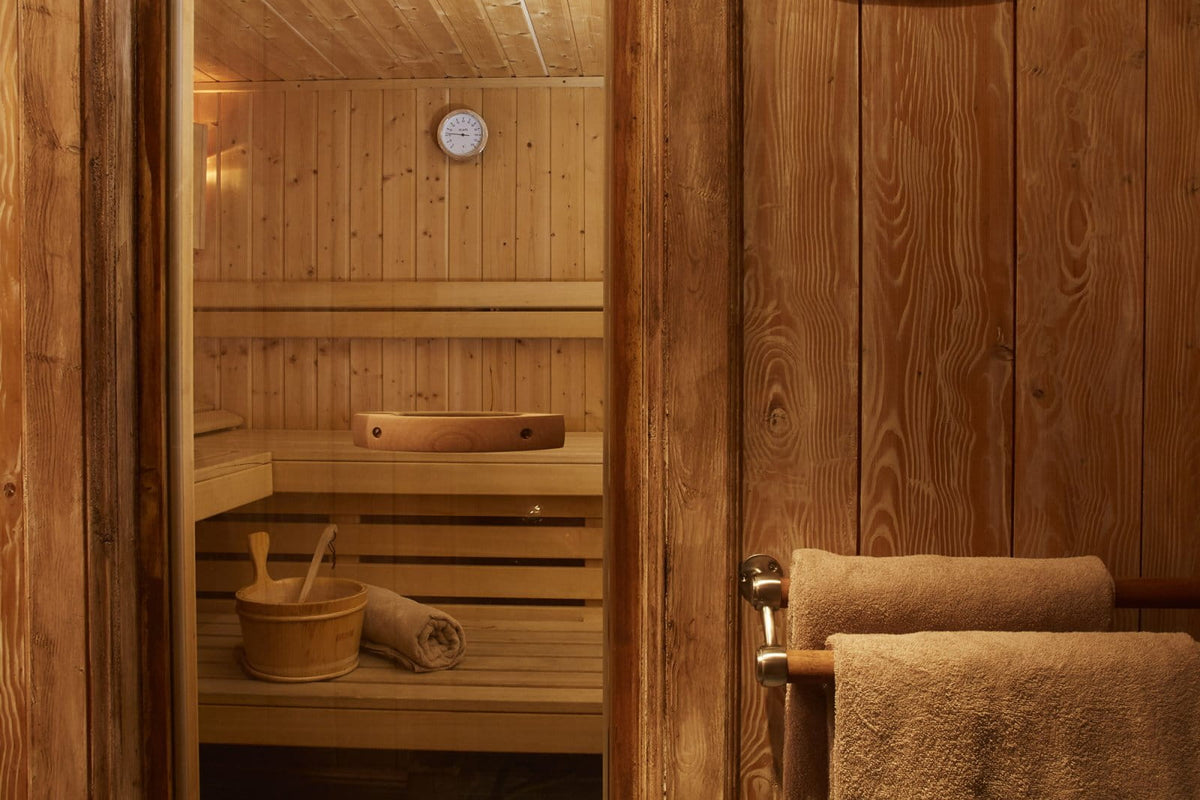 A warm wooden sauna interior with tiered wooden benches, a hanging towel, a wooden bucket and spoon set, and a round wall clock.