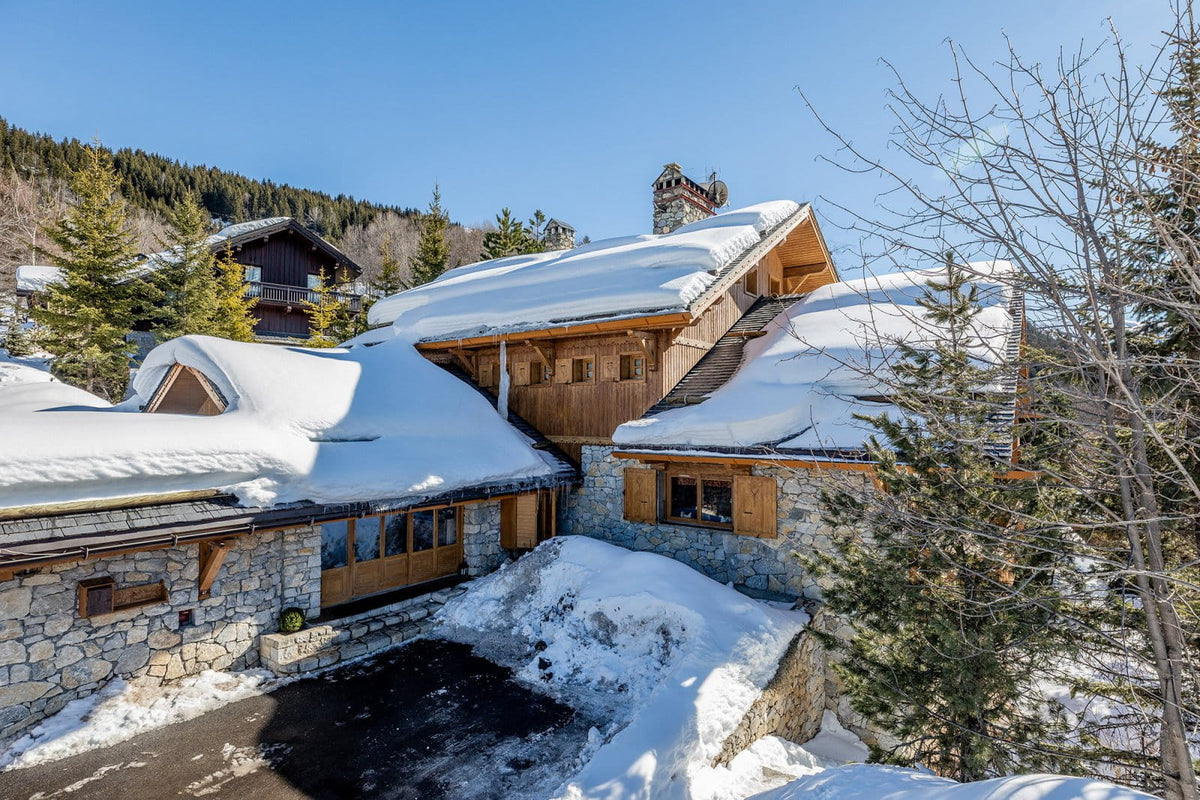 A winter mountain chalet with snow-covered roofs, stone and wood walls, evergreen trees, and a clear blue sky.