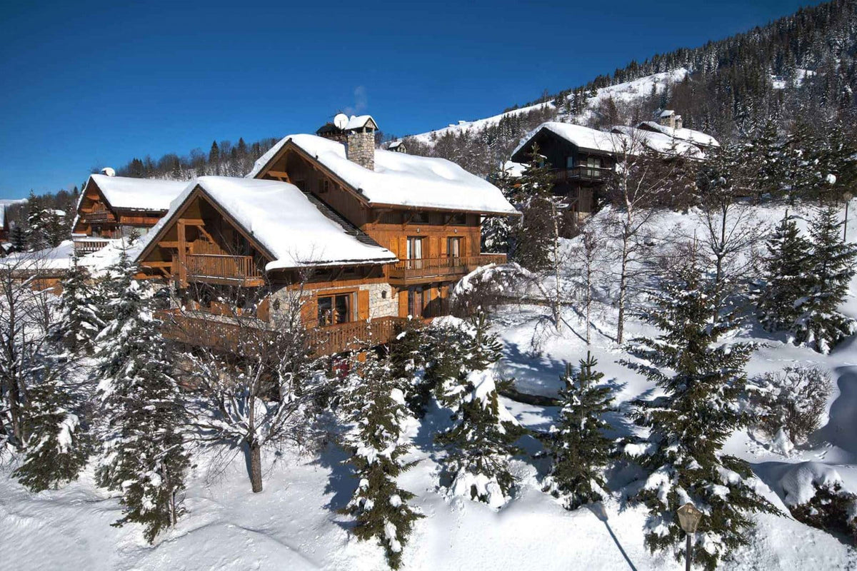 A scenic winter mountain view featuring traditional wooden chalets with snow-covered roofs, surrounded by snow-laden evergreen trees under a clear blue sky.