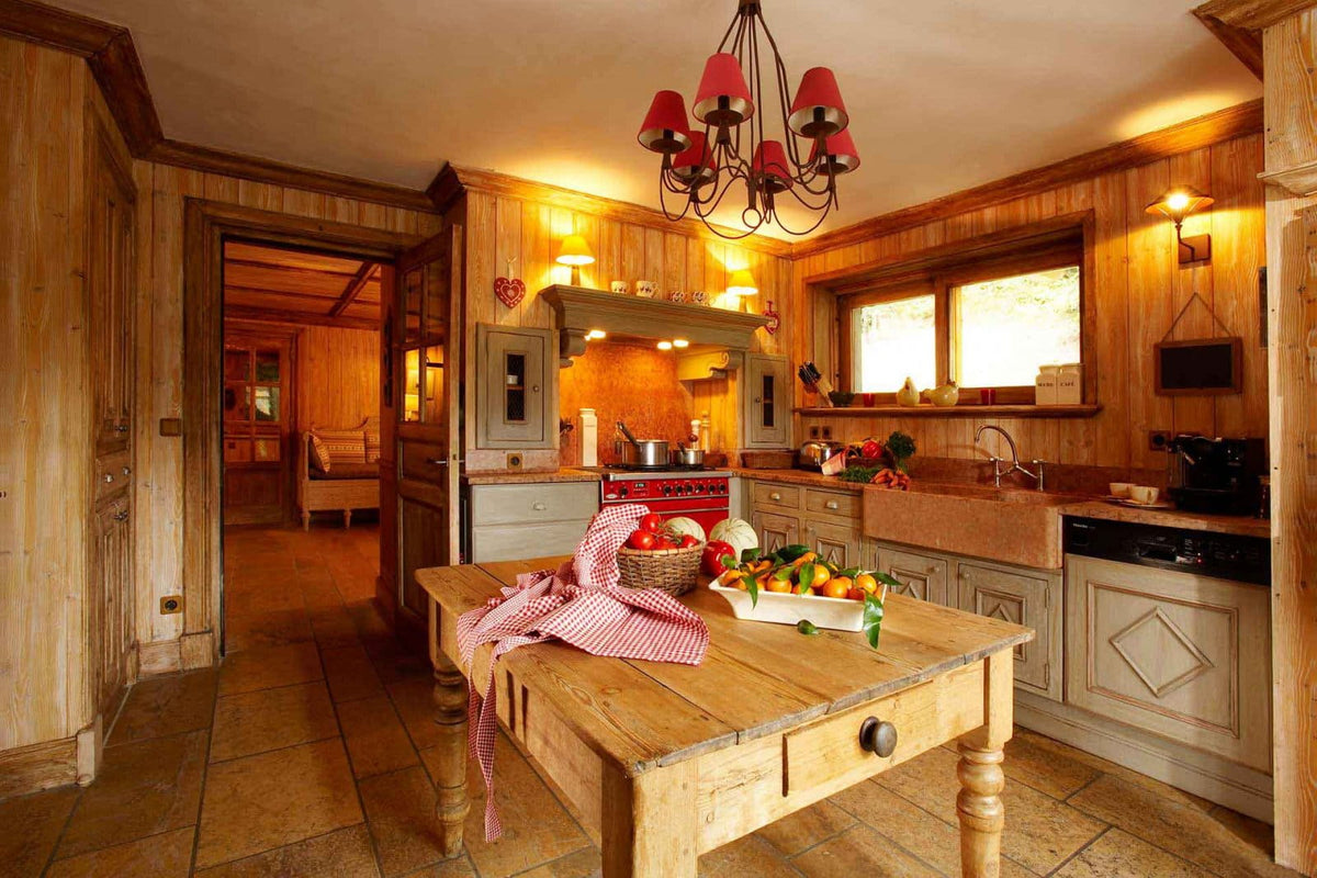 A rustic wooden kitchen interior with a central wooden dining table in the foreground, red pendant lights overhead, wood-paneled walls, tiled floors, and kitchen cabinets in the background.
