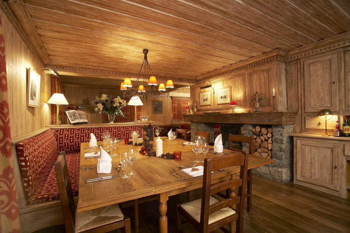 A rustic alpine-style dining room with a wooden table set for six, wooden chairs, a patterned red and beige upholstered banquette, a stone fireplace with wood logs, and warm wood-paneled walls and ceiling.