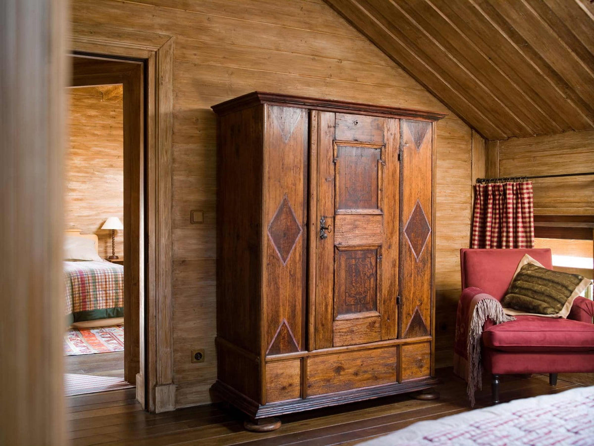 A rustic wooden double-door wardrobe with diamond-shaped inlay details, set in a cozy wood-paneled room next to a red armchair.