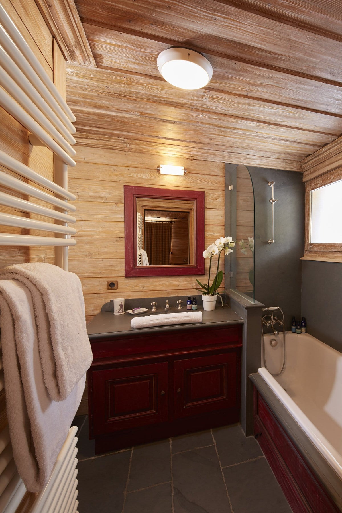 A rustic mountain-style bathroom with wood-paneled walls and ceiling, featuring a dark red wooden vanity with a white sink, a red-framed mirror, a bathtub, and a towel radiator.