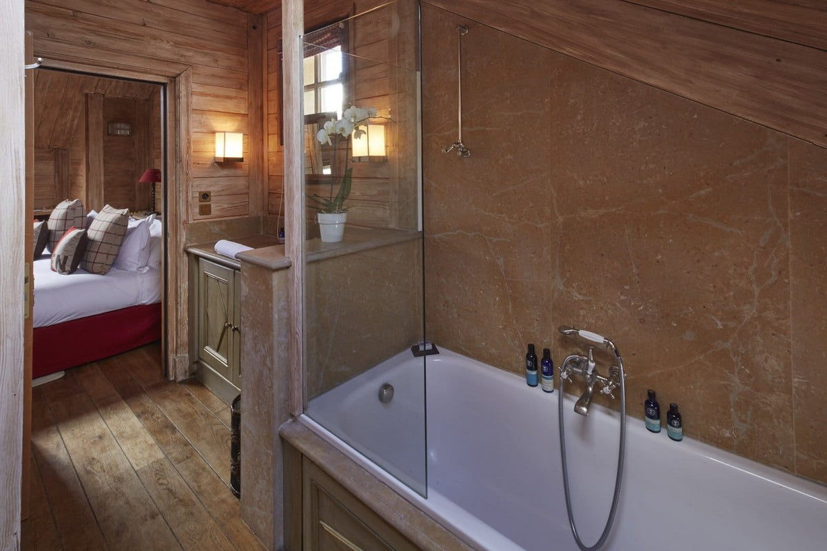 A rustic bathroom interior with a white bathtub set into warm wood-paneled walls, a glass shower screen, and a visible bedroom through an open doorway in the background.
