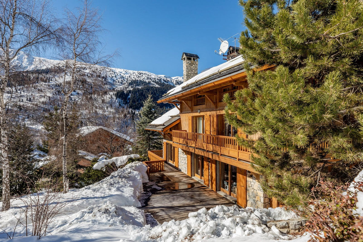 A large wooden chalet mountain house with snow-covered roofs and balconies, surrounded by snow, evergreen trees, and distant snowy mountains under a clear blue sky.