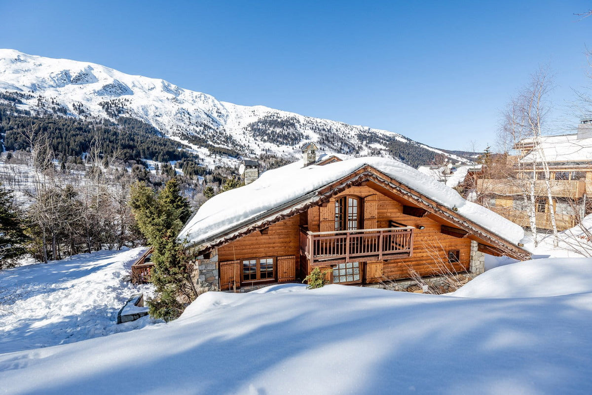 A rustic wooden chalet with snow-covered roofs and balconies, set in a snowy mountain landscape under a clear blue sky.