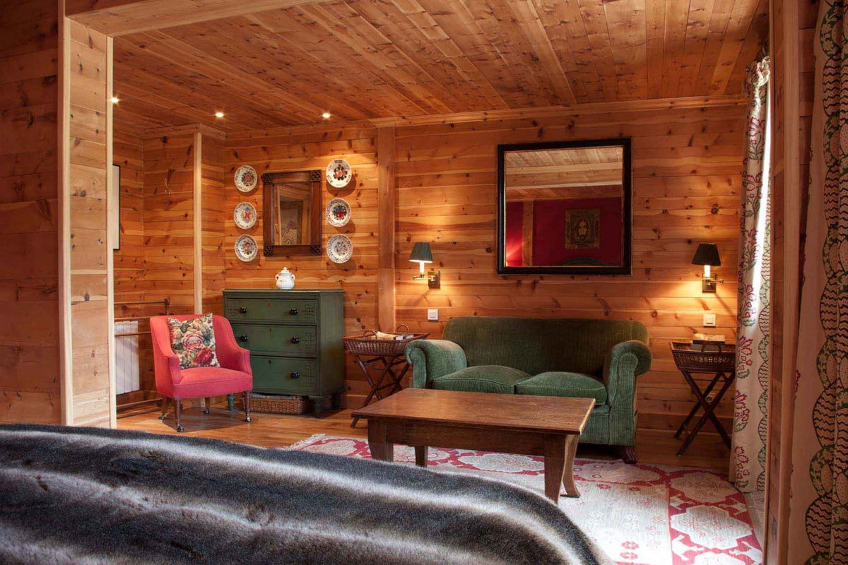 A cozy rustic wood-paneled room with a green sofa, pink armchair, wooden coffee table, and decorative wall plates, viewed from an open doorway.