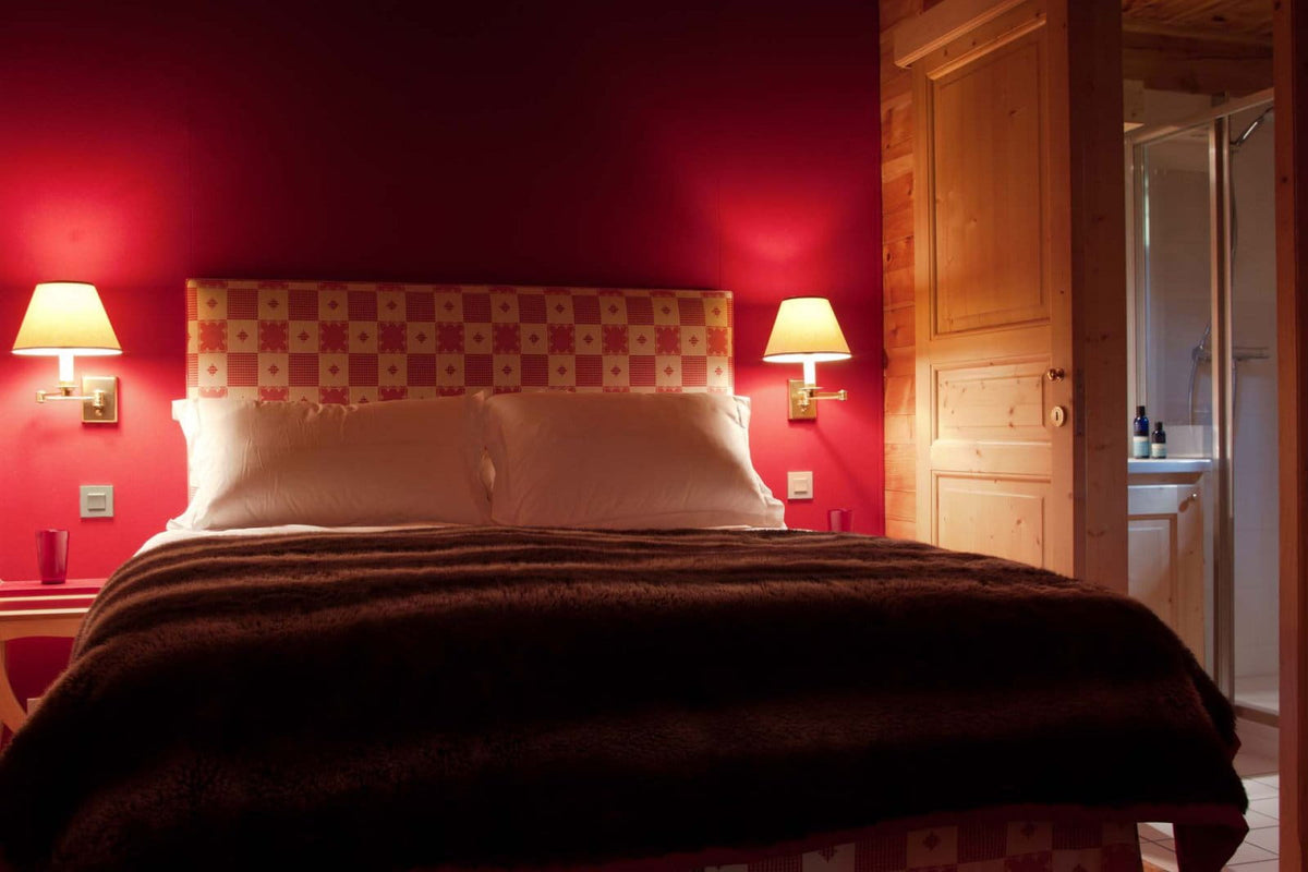 A cozy bedroom scene featuring a bed with a patterned headboard, white pillows, and a dark brown blanket, set against a deep red wall with two lit bedside lamps. A light wood door is open on the right, revealing a glimpse of a bathroom.
