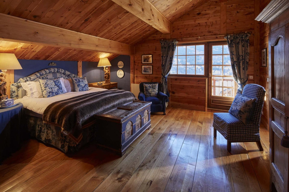 A rustic mountain lodge bedroom with a large bed dressed in blue and white patterned bedding, hardwood floors, wooden walls, and two upholstered chairs near a window.