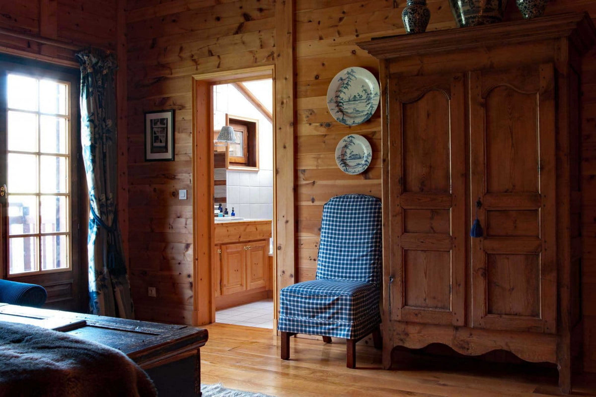 A rustic cabin interior with wood-paneled walls, a large wooden cabinet, a blue-and-white checkered armchair, and a window with curtains on the left.