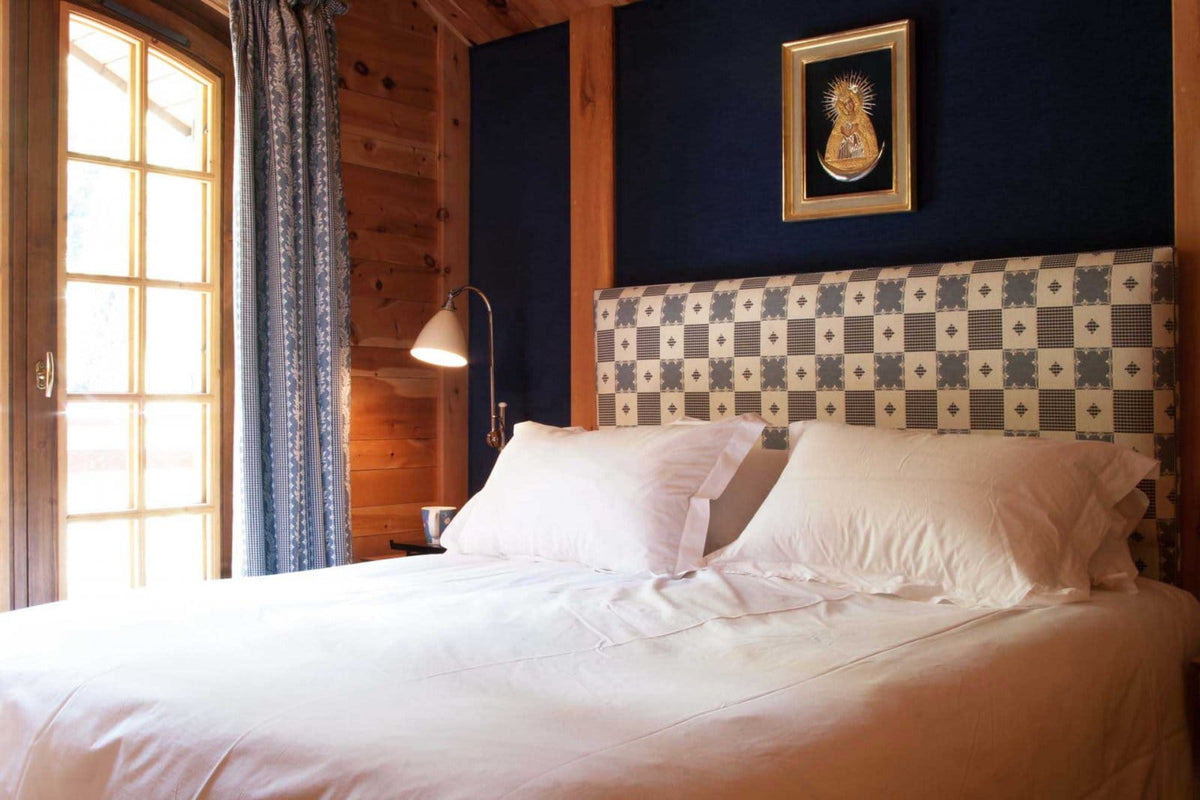 A neatly made bed with white linens in a rustic bedroom featuring wood-paneled walls, a dark blue headboard with a geometric pattern, a window with blue curtains, and a small framed picture on the wall.