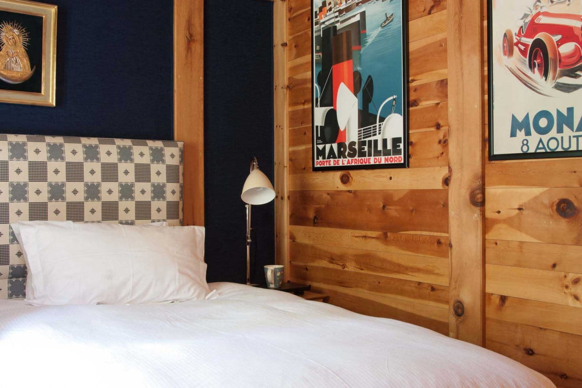 A bed with white bedding and a patterned headboard in a rustic bedroom with wood-paneled walls, a dark blue wall, and vintage car posters.