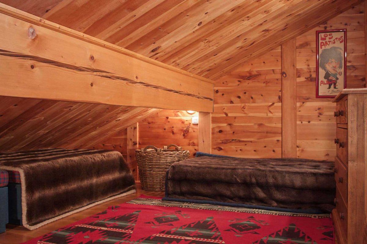 A rustic attic bedroom with two single beds covered in dark brown blankets, set against a wood-paneled slanted ceiling. A red patterned rug covers the floor, and a framed poster hangs on the wall.