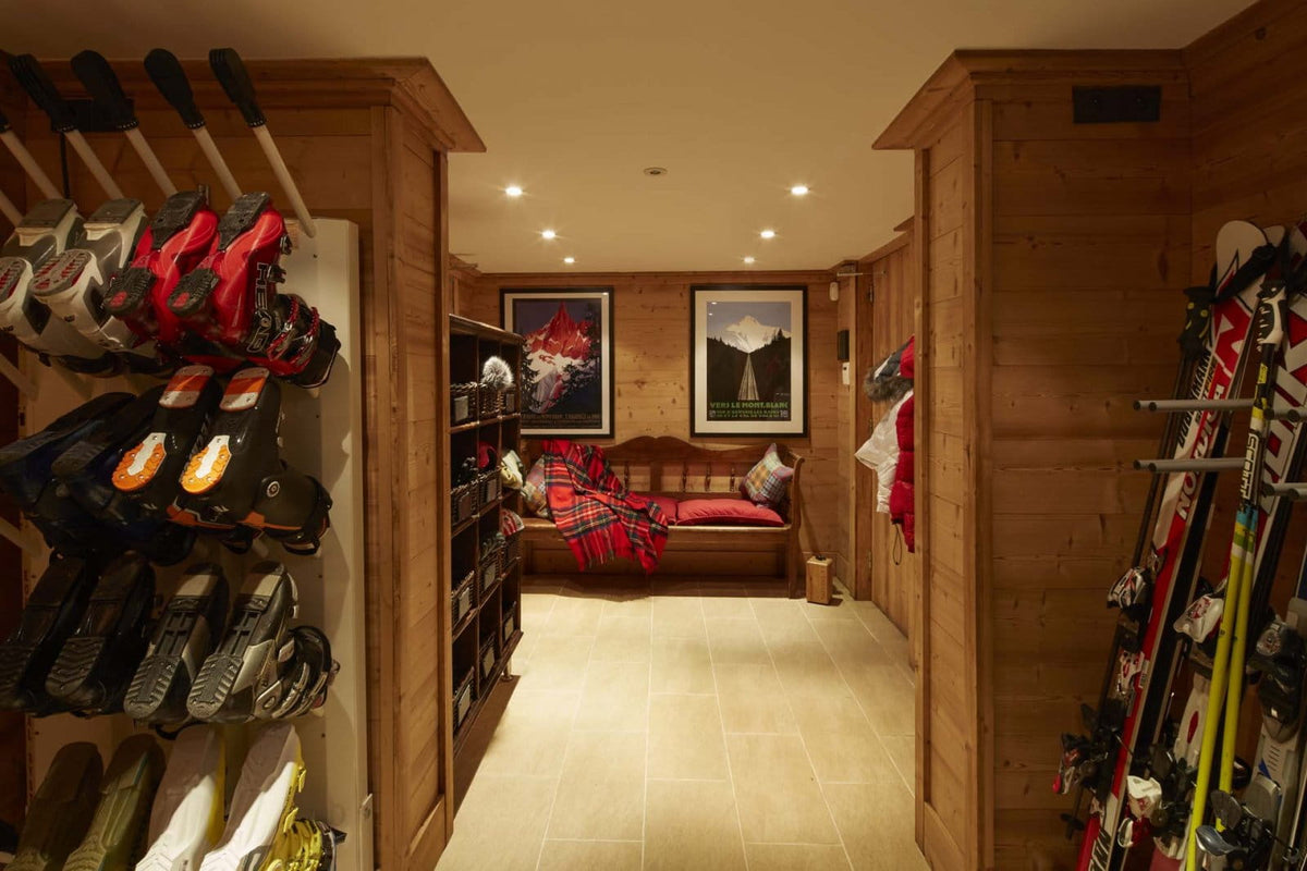 A wooden ski storage room with racks holding skis, snowboards, and ski boots, a bench with a red plaid blanket, framed wall art, and warm recessed lighting.