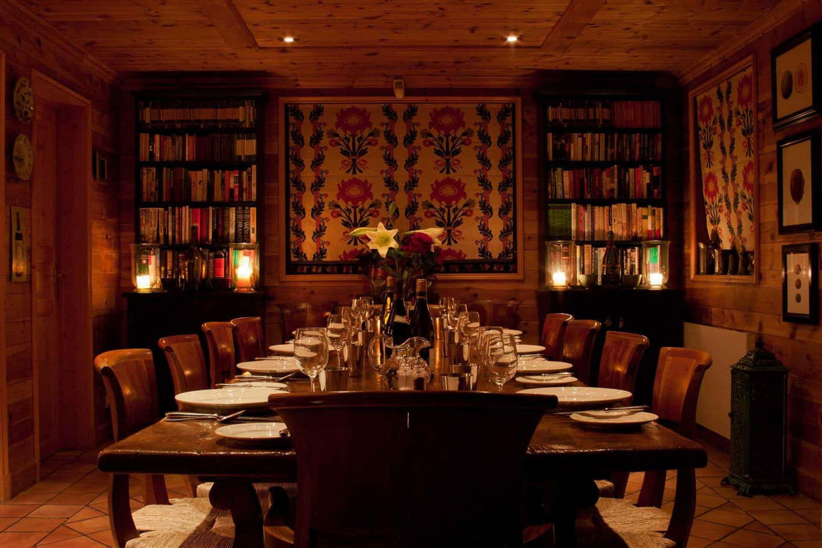 A warmly lit dining room with a long wooden table set for multiple guests, surrounded by dark wooden chairs. The room has wood-paneled walls, tall bookshelves filled with books, and a large floral tapestry hanging on the back wall.