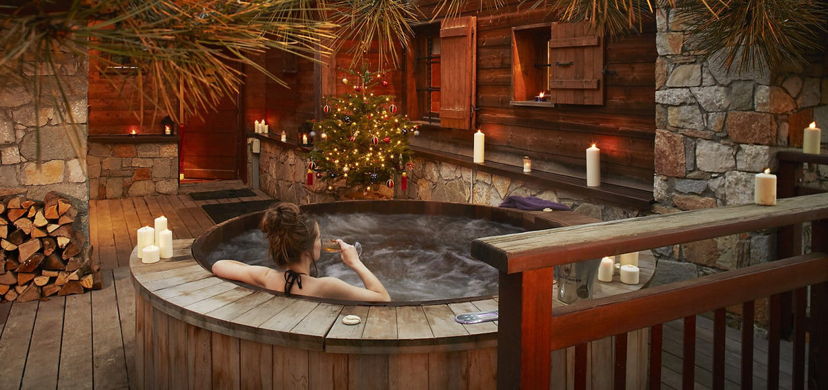 A person relaxes in a round wooden hot spring or outdoor spa tub on a deck, surrounded by candles, logs, and a rustic cabin interior with stone walls and a decorated Christmas tree.