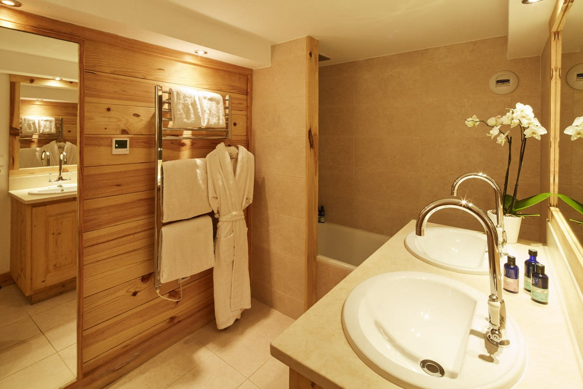 A warm, modern bathroom interior with light wood paneling, two white sinks with chrome faucets, a towel bar with cream robes, and a bathtub in the background.