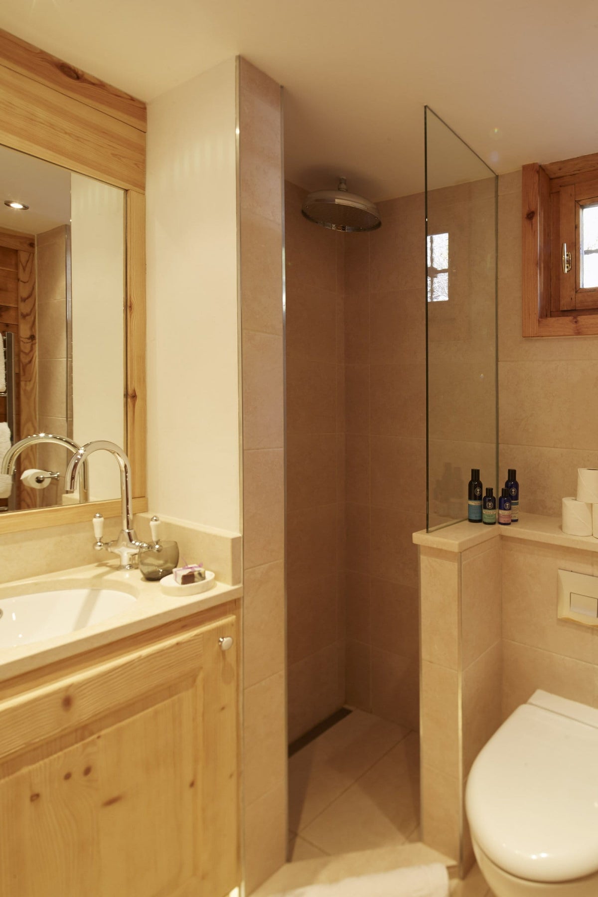 A compact, modern bathroom featuring a light wood vanity with a white sink, a glass shower enclosure, and beige stone walls and floor.