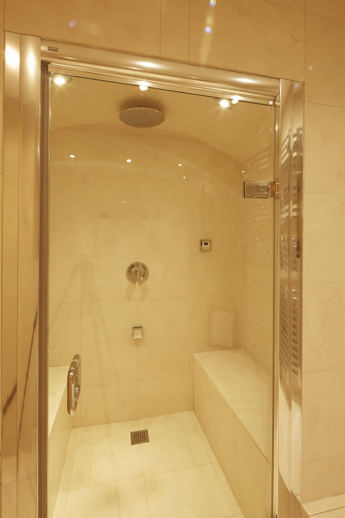A modern walk-in shower enclosure with a clear glass door and chrome frame, set in a tiled bathroom with warm beige walls and floor.
