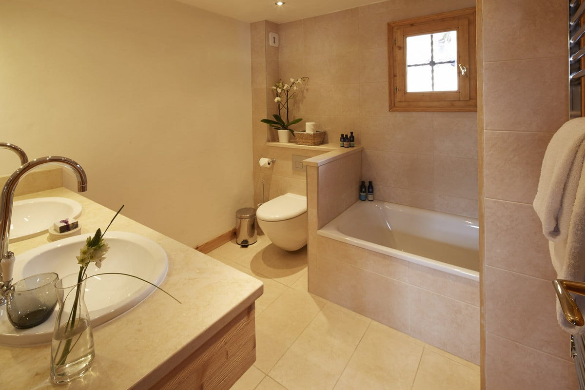 A warm-toned bathroom interior with beige tiled walls and floor, featuring a white bathtub, white toilet, double sinks with chrome faucets, a small wooden window, and a towel on a rack.