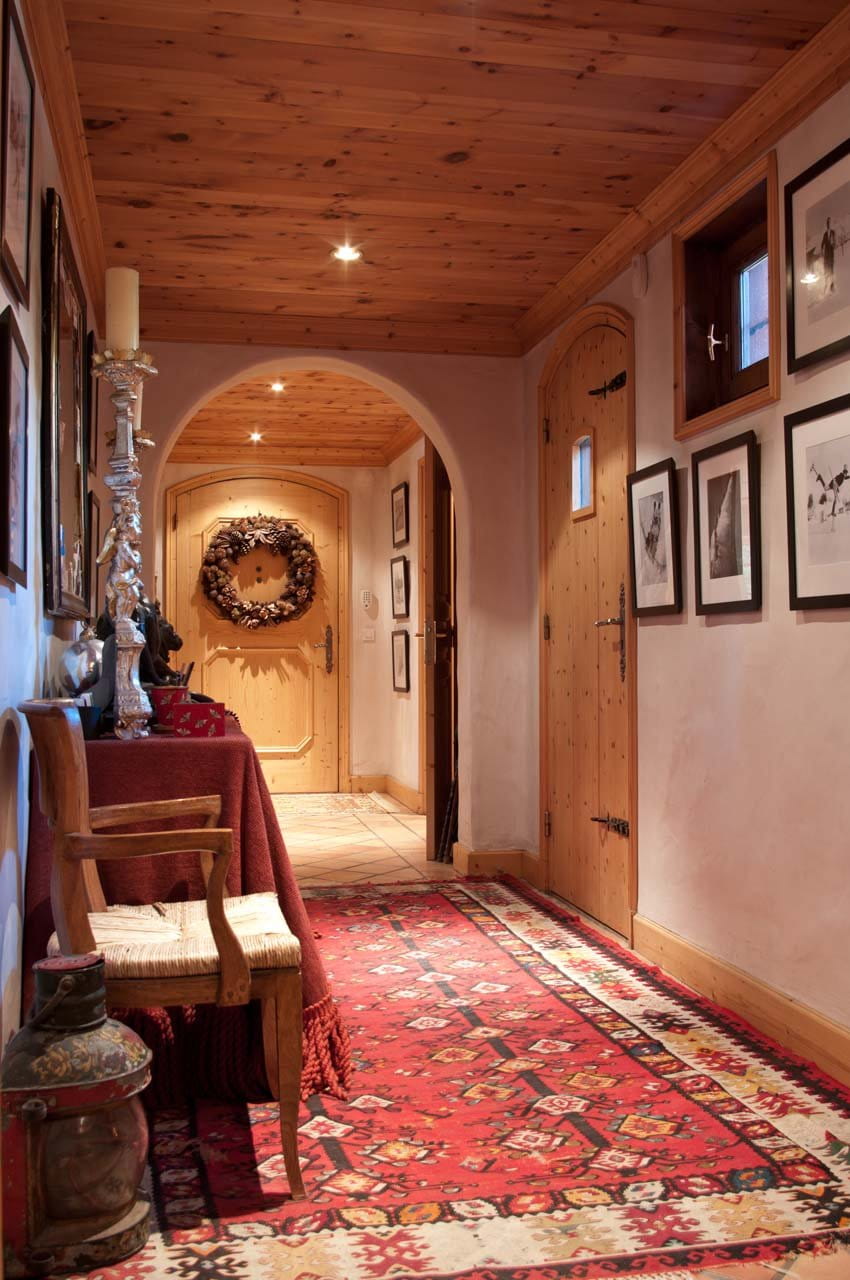 A rustic indoor hallway with a red patterned oriental rug, wooden ceiling, framed wall art, a wooden door, and an arched doorway in the background.