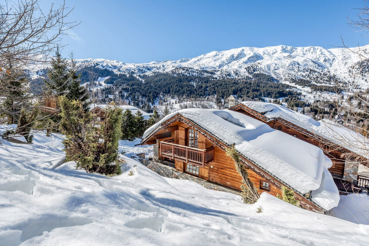 A snowy mountain chalet with wooden walls and snow-covered roofs, set in a deep snow landscape with evergreen trees and snow-capped peaks under a clear blue sky.