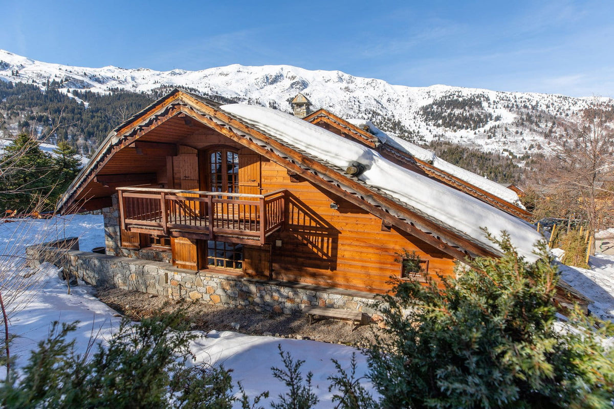 A rustic wooden chalet with snow-covered roofs and a stone base, set in a snowy mountain landscape with pine trees and blue sky.