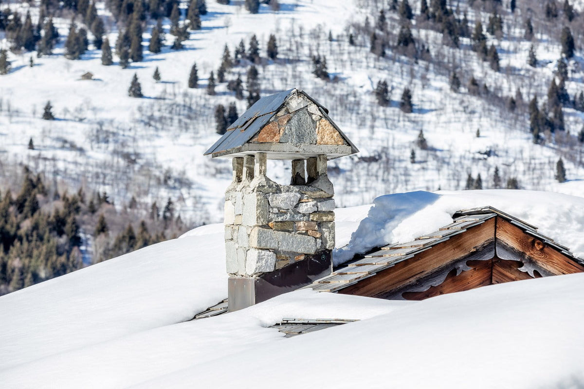 A snow-covered stone chimney with a small metal roof detail, set on a snowy mountain roof with a forested hillside in the background.