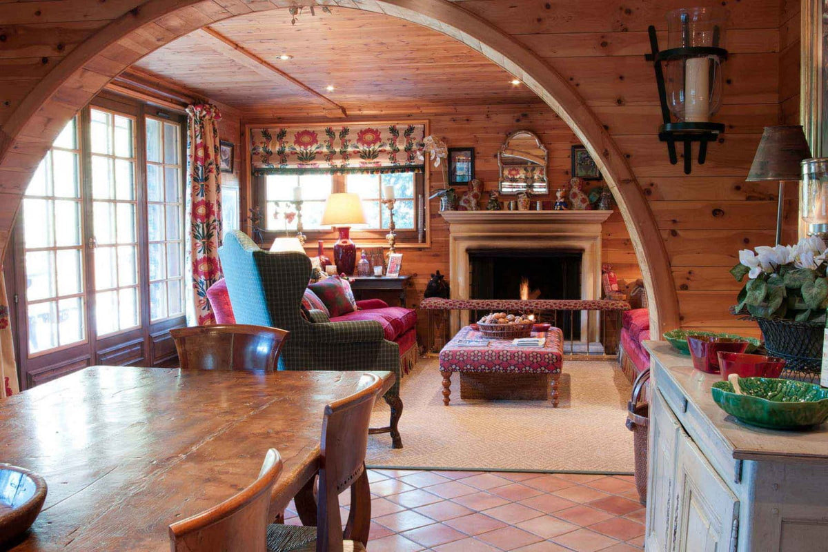 A cozy rustic lodge interior with wood-paneled walls and an arched doorway framing a living room with a fireplace, dining table, chairs, and a coffee table.