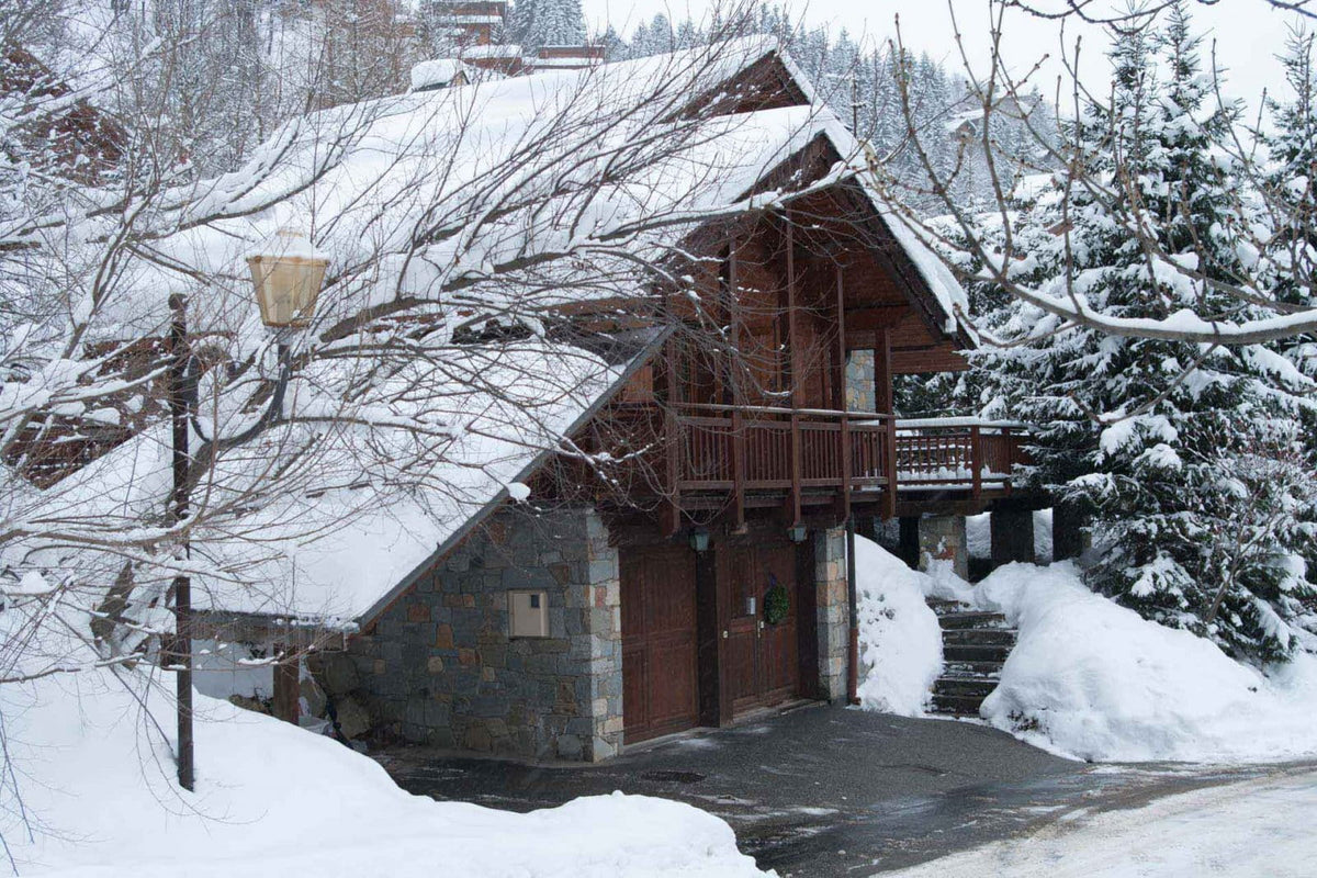 A snow-covered wooden chalet with stone walls and a snow-laden roof, surrounded by evergreen trees and deep snowbanks in a winter landscape.